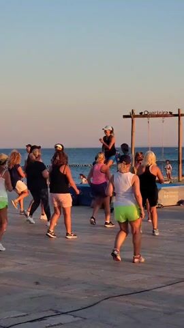 A group of people are dancing on a paved area near a body of water. The sun is setting, casting a warm glow on the scene. A person is standing on stilts in the background. The people are mostly women, dressed in casual, athletic wear. Some are in focus, while others are blurred in the background. The overall mood is festive and energetic.