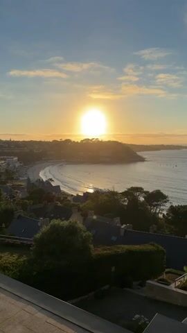 A high-angle, vertical shot captures a serene coastal town at sunrise. The sun, a bright orb, hangs low in the sky, casting a warm, golden glow over the scene. A crescent-shaped beach curves along the coastline, with gentle waves lapping at the shore. The town's buildings, with their dark roofs, are nestled amongst lush green trees, creating a picturesque landscape. The sky is a gradient of blue and orange, dotted with wispy clouds. The foreground features a portion of a balcony or terrace, indicating an elevated viewpoint. The overall atmosphere is peaceful and tranquil, evoking a sense of calm.