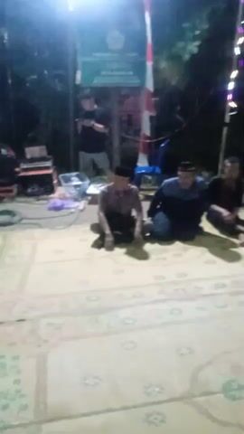A group of men are sitting on a large rug outdoors at night. They are dressed in casual clothing, and some are wearing caps. A sign is visible in the background, along with some audio equipment and lights. The men appear to be conversing or listening to someone speak. The atmosphere seems relaxed and informal.