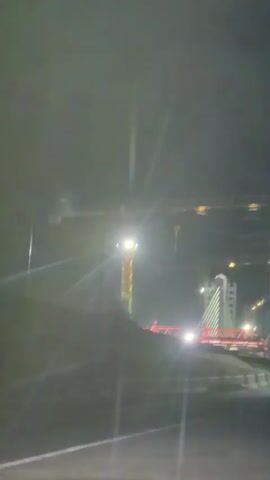 This is a nighttime video showing a construction site. A large yellow crane with a bright light at its center is visible. In the background, a bridge with red supports and white cables is illuminated. There is a tall building under construction to the right of the bridge. The scene is hazy, possibly due to fog or dust, and there are lens flares from the lights. The video appears to be filmed from a moving vehicle, as indicated by the slight blur and the road visible in the foreground.