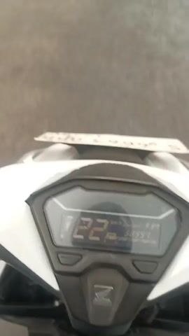 The video shows a first-person perspective from a motorcycle. The digital speedometer displays a speed of 21 km/h and an odometer reading of 30999. The motorcycle is on a paved road in what appears to be a residential or commercial area, with buildings and trees visible on either side. There are other vehicles on the road, including motorcycles and a truck. The camera is slightly tilted upwards.