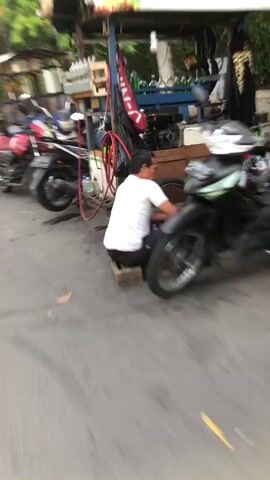 The media file captures a street scene in Pagerwojo, East Java, likely during the day given the lighting. The central focus is a food cart, possibly a mobile beverage vendor, with colorful graphics and images of drinks on its side. The cart appears to be stationary on a paved road.

In the foreground, a person wearing a white t-shirt is interacting with a motorcycle parked beside the food cart. The motorcycle is dark-colored and positioned in a way that suggests it's either parked or being attended to by the person.

The overall image is blurred due to motion, indicating that the recording was likely taken while moving, possibly from another vehicle or by someone walking. This motion blur affects the clarity of details but still conveys the essence of a busy street environment.

In the background, there are glimpses of other stalls or shops, and greenery, suggesting a commercial or populated area. Another person can be seen behind the food cart, possibly the vendor.

The colors are vibrant, especially on the food cart, but the blur makes specific details of the drinks or text difficult to decipher. There's an orange and white striped awning on the food cart. The scene feels lively and typical of a street-side food vending area. There are no obvious indicators of specific weather conditions beyond the presence of daylight. The mood is informal and functional, reflecting a daily commercial activity.