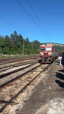 The video shows a red and yellow train covered in graffiti pulling into a train station. The train is traveling on tracks lined with gravel. A person is walking along the platform of the station. The sky is clear and blue. Trees and a hill are in the background.