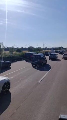 The media file depicts a traffic jam on a highway under a clear blue sky with bright sunlight. The scene is set in Hohenwarsleben, Germany, with several wind turbines visible in the background, indicating a rural or semi-rural setting. The highway appears to have multiple lanes, and numerous vehicles are stopped or moving very slowly.

The primary subjects are the cars and vans stuck in traffic. In the foreground, a white car is partially visible on the left. Further ahead, a dark-colored SUV is prominent in the middle lane, with a license plate that appears to read "WL 3837 Z". To its left, another dark-colored van is in the adjacent lane, and beyond that, another dark SUV or van. Other vehicles, including cars and a white van, are visible further down the highway.

The activity taking place is a traffic standstill, likely due to an accident, construction, or heavy volume of vehicles. The time of day appears to be daytime, given the bright sunlight and clear sky, possibly late morning or afternoon. The weather is sunny and pleasant, with no clouds in sight. The overall emotion conveyed is one of frustration and waiting, typical of a traffic jam. No specific individuals are clearly discernible within the vehicles, but the presence of multiple vehicles suggests a significant number of people are affected by the delay.

The location cues, such as the wind turbines and the visible license plate format, help establish the setting as Germany. The highway infrastructure, including lane markings and barriers, are also visible. There is no discernible text in the media file, other than the license plate on one of the vehicles.