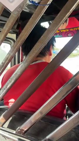 The video offers a first-person perspective from inside a moving vehicle, possibly a moto taxi or a similar open-sided transport, evidenced by the prominent rusty metal bars framing the view. The initial shots (0:00-0:02) look forward, showing the backs of two men; one wears a red shirt, the other a grey or blue shirt. The vehicle's engine sounds are audible.

As the video progresses, the view shifts to the street outside through the vehicle's window. Bright daylight illuminates the scene. A dark SUV is parked on the side of a paved road (0:02-0:03), with multi-story buildings lining the street. A large yellow sign on one building is partially visible, showing the text "BOTICAS FARMA."

The final shot (0:03-0:05) captures another street scene, featuring a distinctive three-wheeled motor vehicle, a moto taxi, painted in green, white, and blue, parked further down the road. Another blue moto taxi is visible in the background. Overhead utility wires and poles are present. The green, white, and blue moto taxi displays the text "POESIDON" and "3213" on its rear, along with a license plate reading "5025 MA." The overall impression is that of a bustling street in a sunny, urban environment.