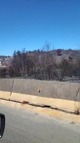 The video shows a view from a moving vehicle traveling on a road with a concrete barrier. On the other side of the barrier, the landscape has been burned. There are charred trees, a black landscape, and a few buildings visible on a hillside in the distance. A power line tower stands in the middle of the landscape. The sky is clear and blue. The camera moves past the burned landscape.