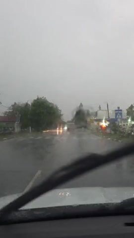The video captures a first-person perspective from inside a vehicle driving through a town during a heavy downpour. The windshield is covered in rain, and the active windshield wipers are visible in the foreground, clearing water to maintain visibility. The sound of intense rain is prominent throughout the video.

The asphalt road ahead is thoroughly wet, with reflections of car headlights creating bright streaks on the surface. Other vehicles, mostly cars and possibly a larger vehicle like a bus or truck in the distance, are seen driving with their headlights on.

On the left side of the road, dense green trees line the street. On the right, various buildings are visible, some with illuminated signs. One prominent sign displays partial Cyrillic text, including "KIPO" and another sign above it reading "УРС СТРОЙ" (URS STROY). A blue square sign indicating a pedestrian crossing is also visible. Briefly, a red traffic light appears on the right side of the road.

The sky is heavily overcast and grey, making the daytime scene appear dim. The overall atmosphere is one of challenging driving conditions due to the adverse weather. No people are directly visible, but the moving vehicles imply drivers. The scene is set in Priseltsi, Bulgaria, experiencing a significant rain event.