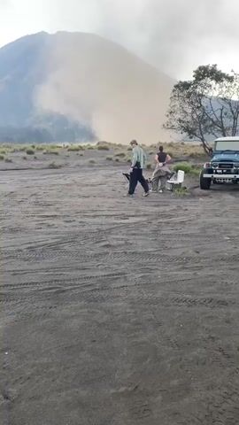 The video shows a sandy expanse with a large, smoky mountain in the background. There are two people, a man and a woman, standing near two folding chairs. To the right of them is a blue and white jeep. The ground is covered with tire tracks. The man is wearing a light-colored shirt and dark pants, and the woman is wearing a dark shirt and light pants. The sky is cloudy. The man moves toward the mountain, carrying one of the chairs.