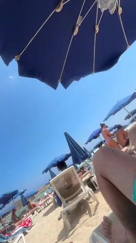 A first-person perspective shot of a crowded beach on a sunny day. The view is angled upwards towards a clear blue sky, with several large blue beach umbrellas dominating the foreground and midground. People are visible relaxing on lounge chairs on the sandy beach, many of them under the umbrellas. Some individuals are wearing swimwear. The overall scene suggests a vacation or leisurely day at the beach.