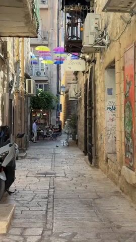 The video shows a narrow cobblestone alleyway lined with stone buildings. Colorful umbrellas are suspended overhead, creating a canopy. There are air conditioning units attached to the buildings. A scooter is parked on the left side of the alley. On the right side, there is a metal gate and some graffiti on the wall. In the distance, there are people sitting at tables outside a shop or cafe, and one person is walking down the alley. A bird is visible on the cobblestones in the middle of the alley.