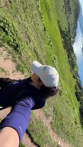 The video begins with a first-person perspective selfie of a person wearing a purple long-sleeved shirt and a white baseball cap with the word "BLAZE" embroidered on it. The person is outdoors on a grassy slope with a body of water visible in the background. The camera then pans to show a group of people hiking on a dirt path through a lush green landscape with rolling hills and a clear blue sky. The hikers are spread out along the path, some walking together, others a bit further apart. In the distance, a mountain peak can be seen. The sun is shining brightly, illuminating the scene.