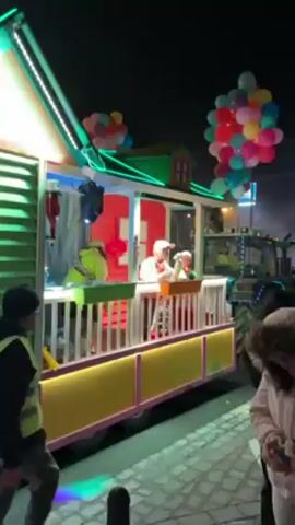 At night, people on the street watch a brightly lit float pass by. The float is shaped like a house with a porch and is pulled by a tractor. Two people on the porch give out balloons to the crowd. The float is decorated with a green roof with neon lights, red walls, and a yellow and pink porch railing. The tractor pulling the float is green. The scene is festive with people gathering to watch the spectacle.