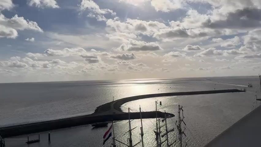 The video shows an aerial view of a harbor entrance with a breakwater curving into the sea. The sun is shining brightly, reflecting off the water. In the foreground, the masts of a sailing ship are visible, with a Dutch flag flying from its stern. Several smaller boats are docked in the harbor. The sky is partly cloudy, with the sun breaking through the clouds. The overall scene depicts a peaceful maritime setting with clear weather conditions.
