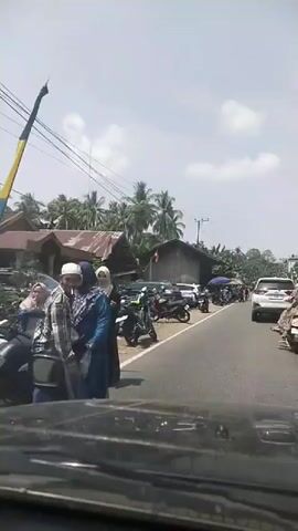 The video begins with a view from inside a vehicle looking forward. In the foreground, the dashboard and part of the windshield are visible. The scene outside shows a road lined with parked motorcycles and some cars. Several buildings, appearing to be houses or small shops with corrugated metal roofs, are situated alongside the road. Palm trees and other tropical vegetation are visible in the background under a bright, cloudy sky.

As the video progresses, the vehicle appears to be moving slowly. On the left side of the road, three women wearing hijabs are standing. One woman in the foreground is wearing a blue and purple patterned outfit, another is in a green hijab and a dark top, and a third woman is further back. More motorcycles are parked along the roadside.

Further down the road, there are more people, some standing and some walking. There are also several stalls or awnings set up, suggesting a market or an event. A white SUV is driving away from the camera down the road, and several other vehicles are visible in the distance. The overall impression is a busy roadside scene in a tropical, possibly rural, setting. The time stamp indicates the video lasts approximately 5 seconds.