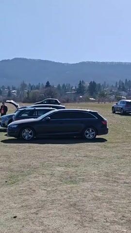 The video depicts an outdoor scene on a clear, sunny day.  In the first shot, several cars are parked on a grassy field, with a backdrop of trees, houses, and a distant mountain range. The camera pans to the right revealing more vehicles, including a tractor.  The next shot features two men standing near a car, their arms crossed and engaged in conversation. Behind them is a plowed field and a mountain range with snow on the peaks.  A final shot shows two SUVs parked near the plowed field, with the mountain view in the background.