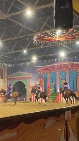 The video shows a group of people on horseback in an indoor arena with a dirt floor. They are dressed in colorful costumes, some appearing to be historical or theatrical. Several individuals are holding bows and arrows, suggesting a performance or competition involving archery from horseback. The background features a stage-like set with columns and painted backdrops that evoke an ancient or classical theme. Decorative lights, including a large, circular light fixture with pink beams, are suspended from the ceiling, indicating an event or show is taking place. The horses are of various colors, and some riders are in mid-action, either riding or preparing to shoot. The overall scene depicts a dynamic performance possibly involving equestrian skills and archery.