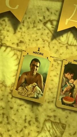 The video displays a garland of yellow paper flags with Cyrillic letters, spelling out "РОЖДЕНИЯ," which translates to "BIRTHDAY" in English. Below the flags, a series of photographs are hung on a string, each in a cardboard frame. These photos appear to document the growth of a baby over its first year. The frames are labeled with numbers and the Russian word "МЕСЯЦА" (MONTHS), indicating the baby's age at each milestone. The first photo shows a man holding a newborn baby. Subsequent photos show the baby with its parents, often with a cake and candles, marking monthly birthdays up to at least 12 months. The background appears to be a textured surface, possibly a blanket or tapestry, with warm lighting that casts shadows. The overall scene suggests a celebratory occasion, likely a baby's first birthday.