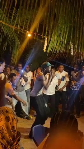 A group of about 15 people dance under palm fronds at night. Many are taking videos on their phones. The ground is dirt. A string of lights hang from the palm fronds. One man dances in the center, wearing a gray tank top and a baseball cap. The light is spotty and unclear.