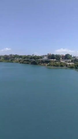 The video shows a view from a bridge with a sidewalk separated from the roadway by a metal railing. The water below the bridge is turquoise in color. On the other side of the water is a city skyline with various buildings including a tall, unfinished gray building with exposed framework. There are trees on the shoreline. On the bridge, there is traffic moving in both directions. The sky is blue with a few scattered clouds. People are walking on the sidewalk of the bridge. A street light and a road sign are also visible.