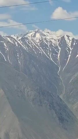 The video shows a view of snow-covered mountains from inside a car. Initially, a red and white road sign is visible in the foreground, with the mountains in the background. The camera then angles upwards towards the car's interior roof, before focusing closely on a person's wrinkled forehead, showing eyebrow hair. Finally, the video returns to the view of the snow-covered mountains against a blue sky with scattered clouds, seen through the car window.