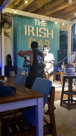 A live music performance is underway at “The Irish Bar” in Gili. A man plays acoustic guitar and sings, backed by a drum set and a bass guitar. A second man dances energetically, moving his arms rhythmically and taking steps side to side. A woman watches from a stool to the dancer’s right. The band performs on a small stage decorated with string lights. The bar includes wooden tables and chairs. A ceiling fan is visible.