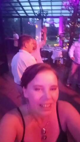 A woman is filming a selfie video. She is wearing a black dress and a necklace. She has brown hair and is smiling and talking. In the background, a band is playing music on a stage. There are people dancing and celebrating. The event appears to be a party or a wedding reception. The lighting is dim and colorful.