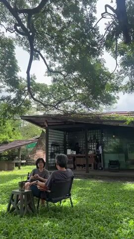 Two individuals, one wearing a grey t-shirt and the other a dark grey t-shirt, are seated outdoors in lawn chairs in a grassy area. To the left of the individuals is a small, grey, foldable stool with a metal cup and a straw on top. Behind them is a building with a slanted roof and glass walls, resembling a cafe or shop. The building has an open-air section where a few people appear to be working or serving customers. The overall environment is lush and green, with large trees providing shade overhead, suggesting a park-like setting. The lighting indicates it is daytime, possibly overcast.