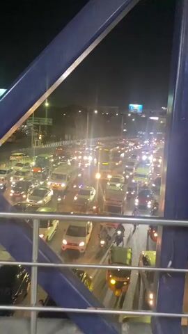 The video shows a heavily congested highway at night. The view is from an elevated pedestrian bridge, looking down at the dense traffic below. Cars, vans, auto-rickshaws, and motorcycles are tightly packed, moving slowly or stopped. The headlights of the vehicles create a bright, illuminated stream of light. Streetlights and billboards are visible in the background. The overall environment is dark, with the bright lights of the vehicles and streetlights dominating the scene. There is a consistent drone of traffic noise.
