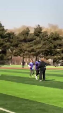 The video shows a soccer field with a group of people dressed in purple jerseys. The people are standing on the field near a white line. Some of them are raising their arms in the air. There is a man dressed in black walking toward the group. The field is surrounded by trees and buildings. The sky is blue and clear. The lighting is bright.