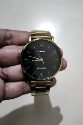 A pair of hands holds a gold-colored Casio watch with a black face. The watch has a metal band with links. The hands are turning the watch to show different angles, demonstrating the design of the watch and its band. The background is a light-colored, possibly textured surface.