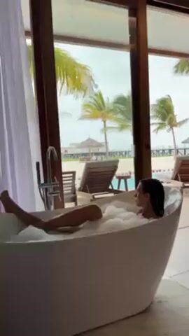 A woman is lying in a white, freestanding bathtub full of bubbles. She is facing the ocean, which is visible through a sliding glass door. There are lounge chairs on a deck outside the door, and palm trees are visible in the distance. The room is well-lit and has a luxurious feel.  The woman is relaxed and appears to be enjoying a bath.
