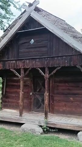 The video shows an outdoor scene at what appears to be a historical village or museum. In the foreground, the side of a dark wooden building with a thatched roof is visible. The building has a porch with wooden pillars. Moving further into the scene, a pathway lined with a wooden fence leads to other structures. Several people, including adults and children, are walking along the path and gathered near one of the buildings. A distinctive wooden birdhouse mounted on a pole stands in a grassy area. The sky is overcast with clouds, and there are trees in the background. The overall impression is of a rustic, traditional outdoor environment.