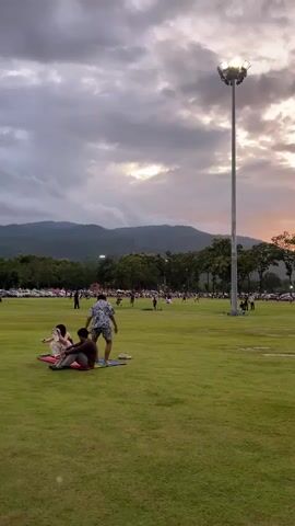 The video shows a wide shot of a large, well-maintained grassy field. In the foreground, four people are gathered on a colorful mat. One person is sitting, another is lying down, and two others are standing. The standing individuals appear to be interacting, with one gesturing with their arms. The field is mostly empty except for these individuals, with a few scattered people further in the background engaged in activities like playing and relaxing. A tall, prominent lamp post with multiple lights stands to the right of the frame, illuminated despite the setting sun. In the background, there is a line of trees and a mountain range. The sky is cloudy, with hints of orange and yellow suggesting it is either sunrise or sunset. There are cars parked along the edge of the field, indicating a public gathering space or park. The overall atmosphere is relaxed and casual, with people enjoying the outdoor space as daylight fades.