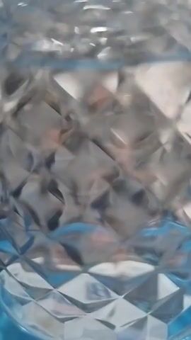A close-up, vertical shot shows a diamond-patterned glass container filled with a clear liquid. The liquid has small ripples and disturbances on its surface, suggesting it is being agitated or poured. The background is blurred, but hints of color, possibly blue, are visible beneath the glass. The lighting is bright, creating reflections on the glass and highlighting the texture of the pattern.