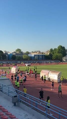 The video shows a running event taking place on a red track within a stadium. A crowd of people are running, many wearing bright yellow or green shirts. Some people are walking near the track. Several white tents or structures with visible logos are situated on the infield. A low stadium seating area with red seats is visible in the foreground. A blue building and trees can be seen in the background under a clear blue sky.