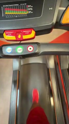 A first-person perspective video shows a person running on a treadmill. The person is wearing red shorts and red sneakers. The treadmill console is visible at the top of the frame with colorful buttons. The person's legs and feet are in motion as they run, and the surrounding environment is blurred due to the movement. The video appears to be recorded from the perspective of the person running.