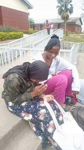 The video shows two young women sitting on concrete steps outdoors. The woman on the left is wearing a dark green camouflage jacket over a floral print dress and a black headscarf. She is looking down with her hands clasped. The woman on the right is wearing a white hooded jacket and bright pink pants. She is looking down at her phone. A white tote bag and a dark backpack are on the steps near them. In the background, there are more people walking and standing near a brick building with a white railing. The environment appears to be a school or institutional campus.