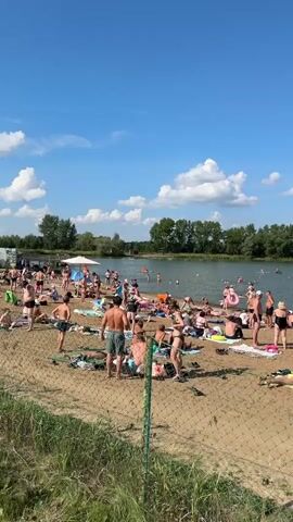 The video shows a crowded beach on a sunny day. Many people are sunbathing on towels on the sand, while others are in the water swimming or wading. There are trees and foliage in the background, and the sky is blue with scattered clouds. Some people are wearing swimwear, and there are beach umbrellas and bags visible. A chain-link fence is in the foreground, partially obscuring the view. The overall scene depicts a lively summer atmosphere at a popular swimming spot.