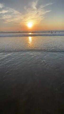 A slow-motion video shows a beach scene during sunset. The sun, a bright orb, is positioned high in the sky, casting a golden glow on the rippling ocean water. Gentle waves roll towards the shore, creating a serene and tranquil atmosphere. Several people are visible in the ocean, some of whom appear to be surfing or swimming. The sky is painted with soft clouds tinged with hues of orange and yellow, further enhancing the picturesque sunset. The foreground of the video consists of dark wet sand, indicating the tide has recently receded. The overall impression is one of peace and natural beauty.
