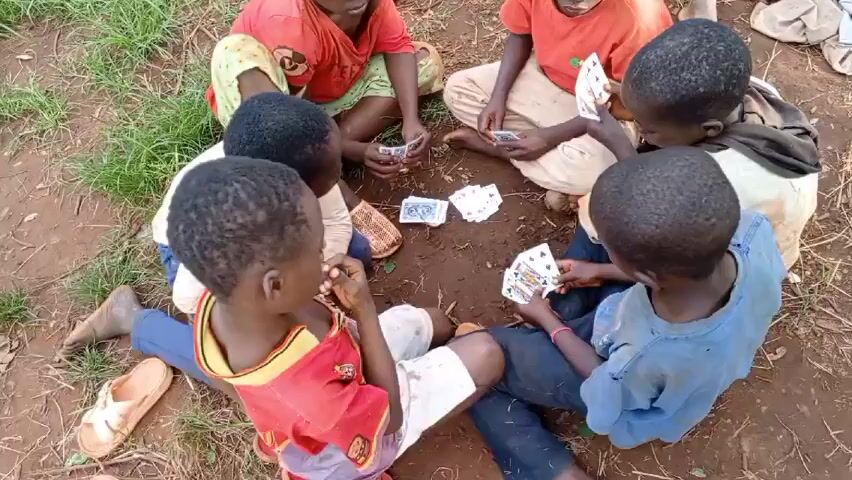 Six children are sitting in a circle on the ground, playing cards. The ground is mostly dirt and small patches of grass. Some of the children are holding cards, while others have cards placed in the middle of the circle. They are all looking at the cards, seemingly engaged in the game. Their clothing is casual and colorful. Some have sandals placed to the side, while others are barefoot. An older person is standing on the periphery of the circle, partially obscuring the scene.