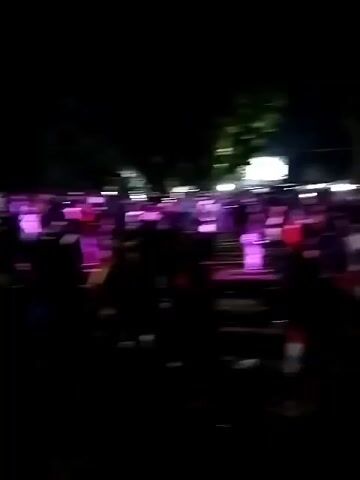 The video shows a nighttime outdoor gathering of people in front of a stage. The stage is illuminated by bright lights, and performers are visible on it, some of whom appear to be singing or playing instruments. A large crowd of people is gathered in front of the stage, many of whom are silhouetted against the lights and appear to be watching the performance. Some individuals in the crowd are holding up their phones, likely to record the event. The environment appears to be somewhat rural or undeveloped, with trees visible in the background. The overall atmosphere suggests a concert or festival.