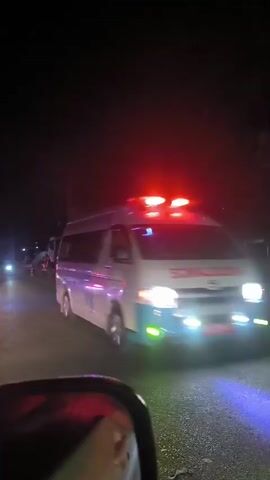 The video opens with a close-up of a white ambulance moving from left to right. The ambulance has "AMBULANCE" written in red capital letters on its side, a blue wavy stripe design including a large "W", and a red shield logo with "119" inside it. The top edge of the ambulance features a blue and white checkered pattern. The scene then shifts to a dark road at night, illuminated by the camera's headlights. In the distance, the red taillights of another vehicle are visible. A person on a motorcycle, also using headlights, passes by the camera's position. The road surface is asphalt, and there is vegetation lining the side of the road. The activity depicts vehicles traveling at night in what appears to be a rural or semi-rural area.
