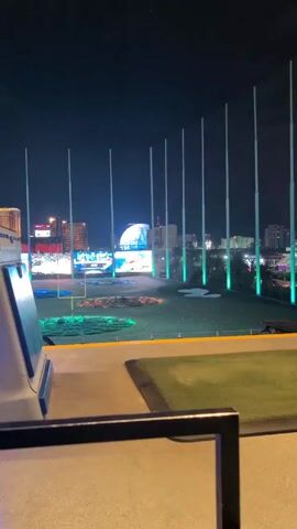 The video shows an outdoor Topgolf location at night, with the Las Vegas cityscape in the background. Several tall poles line the edge of the golf area. Large digital screens display images and advertisements, including a "Hungry for Football?" advertisement with a football and drinks. The MSG Sphere is visible in the distance behind the screens.