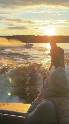 The video shows a lake with a sunset in the background.  A person is riding a jet ski on the water, creating waves.  The video is taken from a boat with two people in it.  One person is standing up looking at the jet ski, while the other is sitting down wearing a hooded sweatshirt. The horizon has a line of trees on the far shore.  The setting sun reflects off the water.