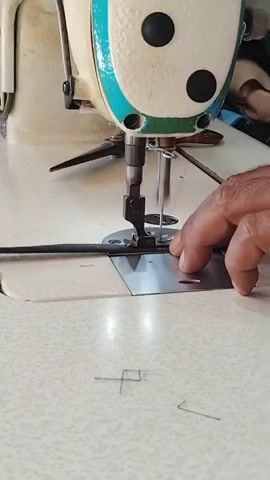 This is a close-up, eye-level shot of a person's hands operating a sewing machine. The sewing machine is cream-colored with turquoise accents and appears to be industrial. A black cord or piping is being fed under the presser foot and needle. The person's right hand is guiding the piping, while their left hand is near the presser foot, likely controlling the fabric or the piping. The tabletop of the sewing machine is light-colored and speckled. There are some loose threads on the tabletop, indicating ongoing work. The lighting is bright, illuminating the details of the machine and the hands. The shot is focused on the action of sewing.