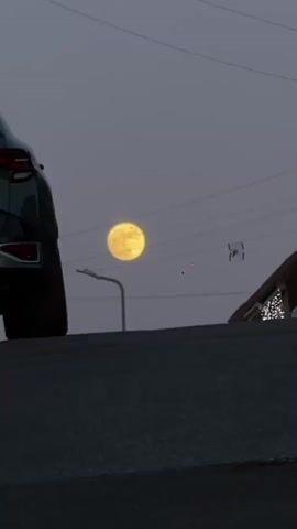 The video captures a low-angle view of a residential street at dusk or dawn. A dark-colored SUV is parked on the left, partially obscuring the view. In the background, a large, yellow full moon dominates the sky. Power lines stretch across the sky, and a street lamp is visible between the car and the moon. A house with lights outlining the roof can be seen on the right side of the frame. The foreground is a dark asphalt road, slightly elevated.