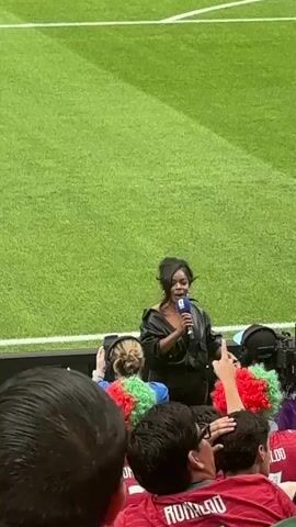 This media file captures a live event, likely a sports match or a pre-game ceremony, taking place in a stadium with a vibrant green grass field. In the foreground, a diverse crowd is visible, many wearing colorful wigs and sporting jerseys, suggesting an atmosphere of excitement and fan engagement. One jersey prominently displays the name "RONALDO" and the number "7" in white lettering against a red background.

The central figure is a woman dressed in black, holding a microphone and appearing to address the crowd or be involved in a broadcast. Her posture and the presence of a microphone indicate she is likely a host, commentator, or performer. Behind her, equipment suggestive of media production, such as cameras and audio gear, is present.

The lighting and the clean, well-maintained field suggest the event is taking place during daylight hours, possibly in the afternoon or early evening. The overall mood appears energetic and festive, fueled by the enthusiastic spectators. While specific weather conditions aren't explicitly shown, the clear visibility and outdoor setting imply fair weather. The visual cues, particularly the stadium environment and the sports-related attire of the spectators, strongly point towards a sporting event taking place in a stadium setting. The mention of "Houston, United States of America" as a location context further narrows down the possibilities to a major sporting venue within that city.