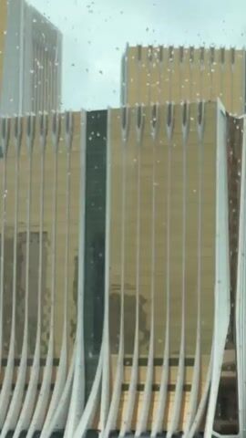 The video was taken from inside a moving vehicle, looking out the window. Raindrops are visible on the window. Through the window, we see a cityscape with several tall buildings. One building is covered in gold and silver vertical lines. We see the Galaxy Arena with a large screen displaying an advertisement. We also see the Venetian hotel in the background. There is some greenery visible between the buildings. The camera pans to the right to reveal more of the buildings. There are muffled conversations in the background.