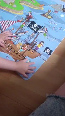The video shows a young child's hands interacting with a large floor puzzle depicting a pirate ship scene. The puzzle is on a wooden floor. The child is placing or adjusting a puzzle piece near the bottom right of the main pirate ship image. The puzzle includes pirate flags, characters on the ship, and water with some sea creatures. The child is wearing denim shorts. The camera is positioned above and to the right of the child, focusing on the puzzle and the child's hands.