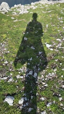 The video shows a shadow of a person standing on a grassy, rocky area. As the camera tilts up, a scenic view of a body of water surrounded by mountains is revealed. The town is nestled along the water's edge. The sky is partly cloudy.